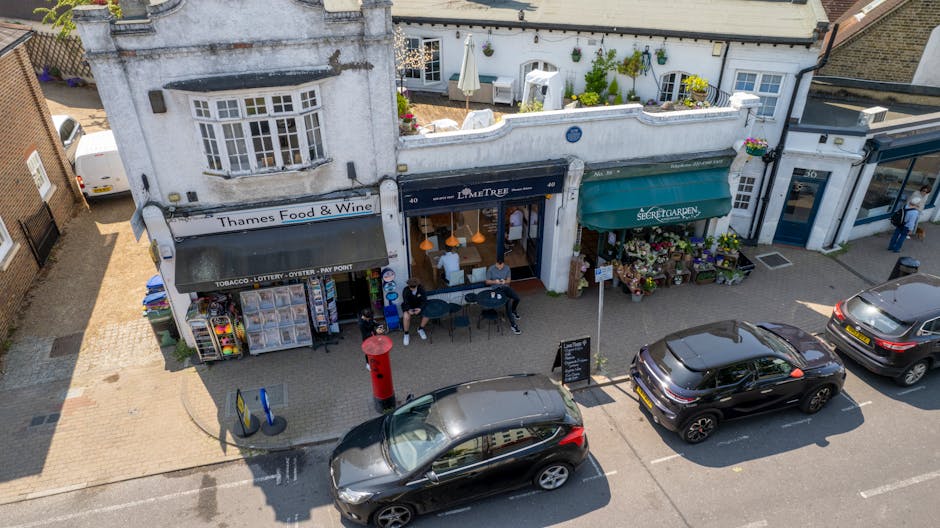 An aerial view of a small commercial street featuring a florist shop with a green awning labeled 'The Secret Garden,' displaying a vibrant floral arrangement outside. Next to it, a café has outdoor s