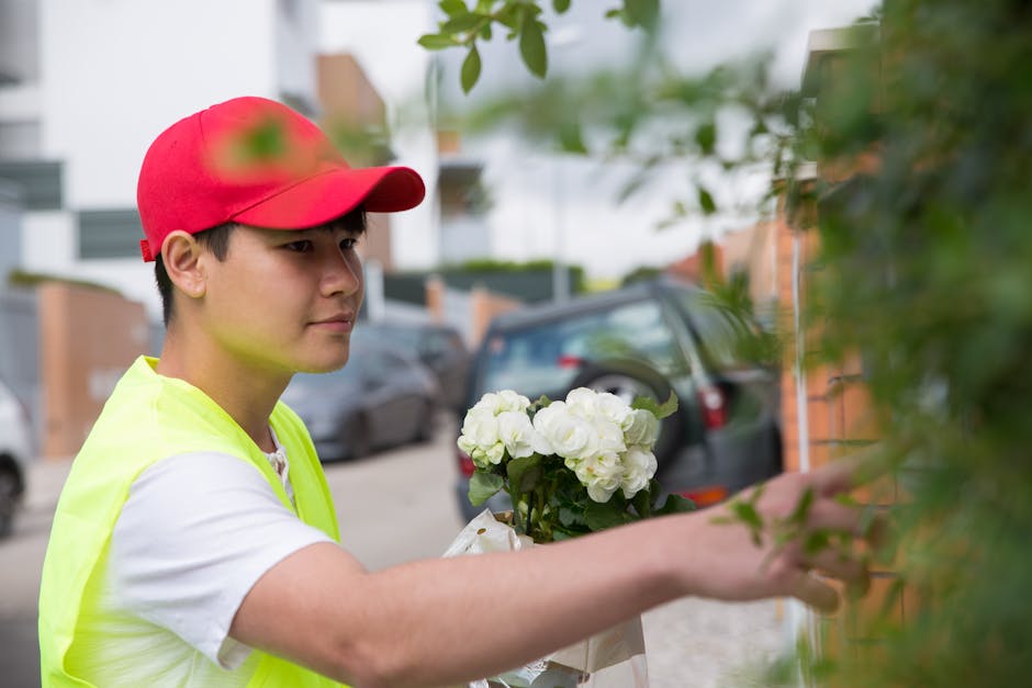 A young male florist with light skin, wearing a red cap and a high-visibility yellow vest over a white T-shirt, is delivering a floral gift outside a residential building. He is holding a bouquet of f